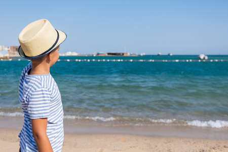 Happy Stylish Boy With Hat Enjoys Life on Summer Beach and Looking Into Distance to Sea Summer Vacation Concept Copy Spaceの写真素材