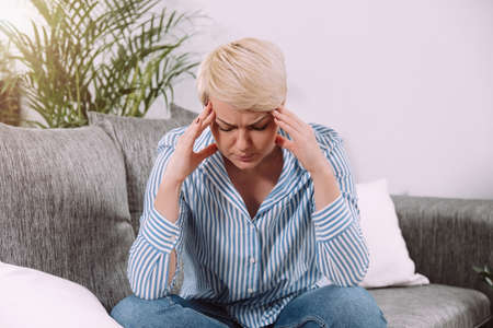 Woman sitting on couch at home with closed eyes holding head with hands and suffering from migraine or headache pain, stress or depression.の写真素材