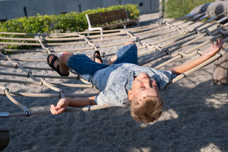 Cheerful blond boy smiling and lying on a rope swing in public park.の写真素材