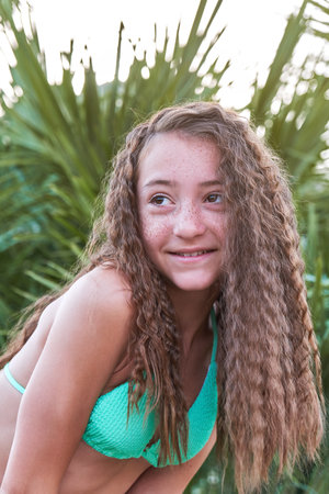 Teenage girl with freckles and curly long hair in bathing suit poses against backdrop of a palm treeの写真素材