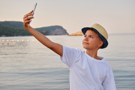 Boy in a hat takes a selfie portrait near the sea. Happy guy enjoying summer holidays at beachの写真素材