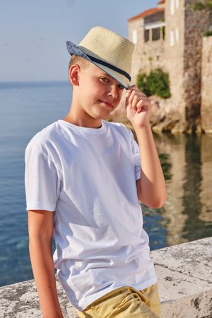 Portrait of a teenager boy in a white t-shirt and straw hat sitting near the sea.の写真素材