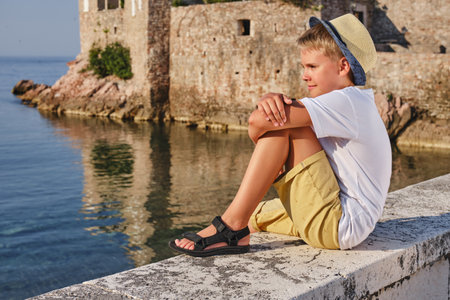 Portrait of a teenager boy in a white t-shirt and straw hat sitting near the sea.の写真素材