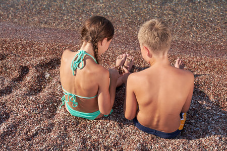 Friends boy and girl are sitting on shore of clean transparent sea and playing with small pebbles.の写真素材