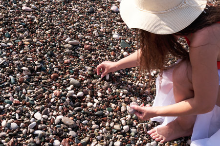 A young woman in a pareo and a sun hat collects pebbles while walking on the beach.の写真素材