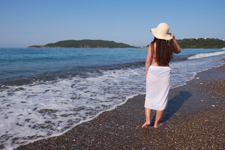 Young brunette woman with long hair in a white pareo and hat walks along the beach.の写真素材