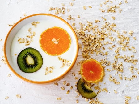 A plate with milk porridge, yogurt, kiwi and orange, with dry oatmeal on a white textured...