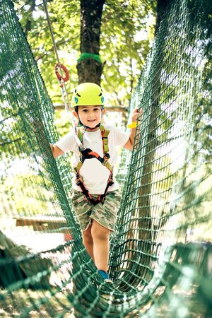 Happy little boy passing the cable route high among trees, climbing and playing, extreme sport in adventure parkの写真素材