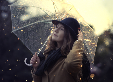 Young woman dreamy mood under rain with illuminated transparent umbrella. Wearing wool coat and stylish hatの写真素材