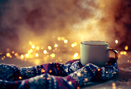 Cozy atmosphere with winter coffee or tea mug with steam covered with wool scarf over bokeh lightsの写真素材