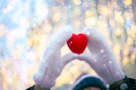 Woman hands in white knitted mittens with a red heart on a snow background.の写真素材