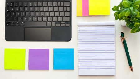 A notebook with blank pages, a laptop, colored sticky notes, paper clips, and a pen on a white table...