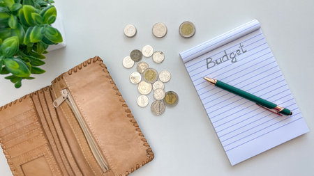 Coins, an open leather wallet, and a notebook with the word "Budget" written on it on a white table. Concept for personal finance management, spending control, and budgeting.の写真素材