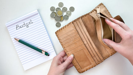 An open leather wallet in hand, with coins and a notebook labeled Budget lying nearby. Conceptually,...