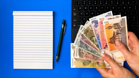 A top view of hands holding Cambodian banknotes next to a laptop, notepad, and pen on a blue background.の写真素材