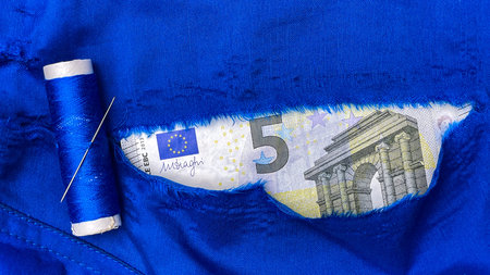 A five-euro note is visible through a tear in the blue fabric next to a needle and thread....