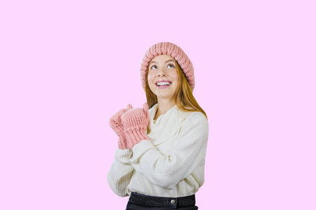 Attractive red-haired girl in a warm hat and mittens looking at the camera against an isolated pink backgroundの写真素材