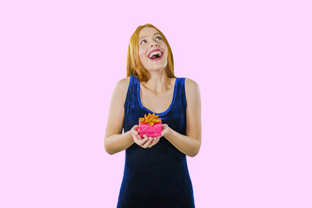 A beautiful red-haired girl in an evening blue velvet dress holds a small box with a gift and presses herself enthusiastically with a scream, smiles, opens her mouth and looks sideways, raising her head against an isolated backgroundの写真素材