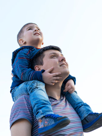 Small boy is sitting on his father's shoulders and pretending a plane during walk in the field