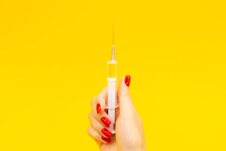A woman's hand with a red manicure holds a syringe on a yellow background. The concept of medicine