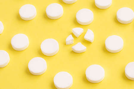 Round white medical pills arranged and one of them is crushed into four parts on yellow background.の写真素材