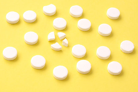 Round white medical pills arranged and one of them is crushed into four parts on yellow background.