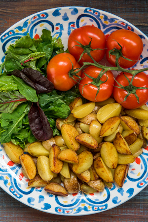 Fried baby potatoes, green salad and fresh tomatoes on a big plate in rustic style.の写真素材