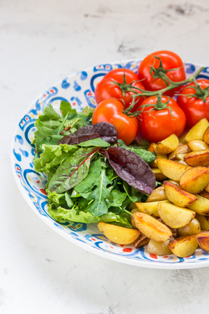Fried baby potatoes, green salad and fresh tomatoes on a big plate in rustic style.の写真素材