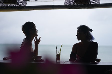 silhouette of 2 asian women relaxing, drinking and talking at beach bar in summer dayの写真素材