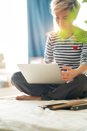 Beautiful Asian woman sitting on bed & working from home on laptopの写真素材