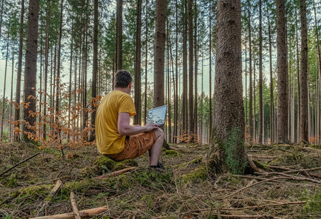 Enjoying work on the fresh air, a man works with his laptop in a natural looking green forest.