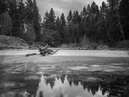 Winter landscape view over a frozen lake with parts of water, which is reflecting trees of the surrounding forest.の写真素材