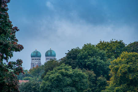View at the tops of the both popular towers of the Frauenkirche in Munich, bavaria over green and autumn colored treetops.の写真素材