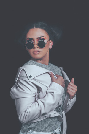 Fashion Girl Posing, wearing Urban Hipster Style, Sweet Pastel Colors, Trendy Sunglasses, Pink Jacket, Dark Background.の写真素材