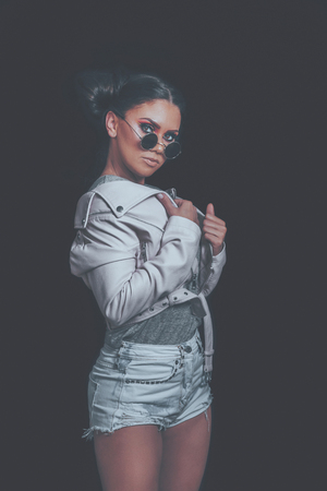 Fashion Girl Posing, wearing Urban Hipster Style, Sweet Pastel Colors, Trendy Sunglasses, Pink Jacket, Dark Background.の写真素材