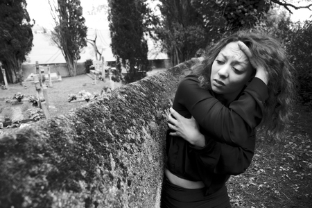 Sad young woman  She is really depressed  She wears black clothes  She stands along the wall of a cemetery  Her pain is very hard の写真素材
