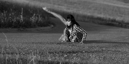 Dancer on a country road. She wears a checked jacket. She sits on the asphalt and she lifts up one leg. She is barefoot. This image is a black and white photography.の写真素材