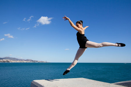 Beautiful classic dancer jumping over a big stone block. This young woman wears dark clothes, black shoes and white tights. In the background, the Mediterranean Sea.の写真素材