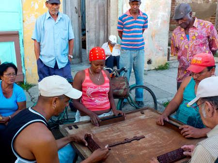 CAMAGUEY, CUBA - DECEMBER 2: people look at three men and a woman Who wooden play dominoes in the street, we december 2, 2014 in Camaguey, Cubaのeditorial素材