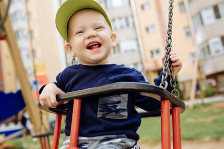 Funny and handsome boy rejoices, rides on a childrens swing, plays on the playground