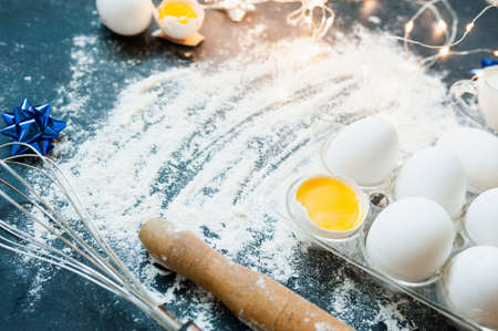 white eggs, a wooden rolling pin, a metal whisk, a glass of milk and festive bows and garlands on a...