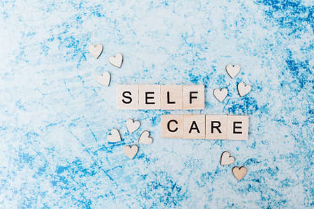 self care lettering in square wooden letters with small wooden hearts on a blue and gray concrete...