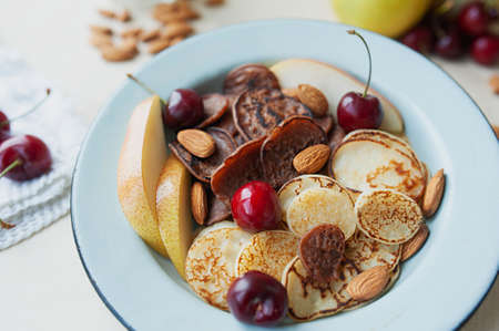 tiny pancakes with pears, cherries, nuts in metal plate against the background of kitchen table with fruit. trending food cereal pancakes. bright and beautiful breakfastの写真素材
