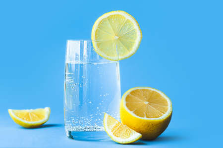 translucent glass of water with gases and bubbles, with a round slice of lemon on a glass and a whole lemon on blue background. Refreshment in the heat. Healthy lifestyle and nutritionの写真素材
