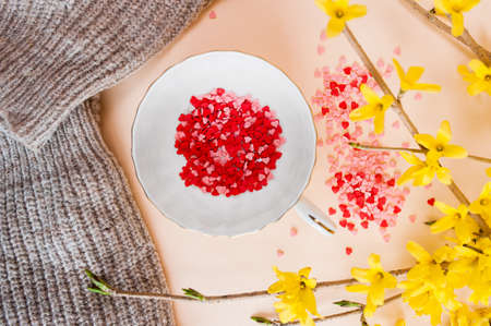 spring tea party. small white cup filled with pink and red spangles stars and saucer on a light background with spring yellow forsythia flowers. Flat lay, top view, Verticallyの写真素材