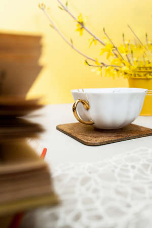 white porcelain Cup with a gold handle on a wooden stand on a light table with old books and a bouquet of spring flowers in a small bucket. spring tea partyの写真素材