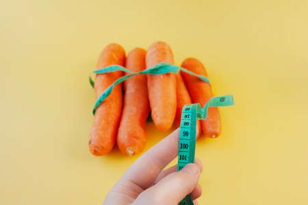 hand holding a centimeter long ribbon on a background of fresh orange peeled large carrots on a yellow background. weight control on quarantine and isolationの写真素材