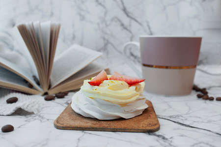 Homemade beautiful Breakfast. White dessert of meringue, cream and fresh strawberries, hot drink mug, open book and coffee beans on white marble surfaceの写真素材