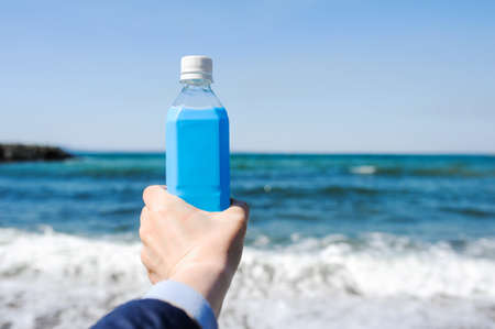 blue plastic bottle in a light-skinned hand against the background of a blue raging sea with foam.の写真素材
