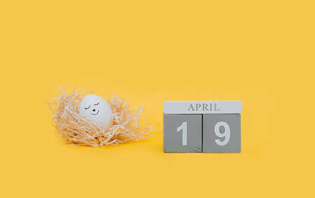 cute white Easter egg with a black marker drawn face sleep in a paper nest made of pressed paper on a clear yellow background and gray wooden cubes with the date of Easter April 19の写真素材